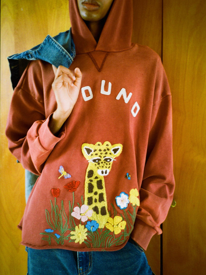 A person wears the FOUND Embroidered Wildlife Hoodie in red, featuring "FOUND" text and leopard, flower, and butterfly embroidery, holding a denim jacket. Made from vintage French-terry cotton for comfort and style.