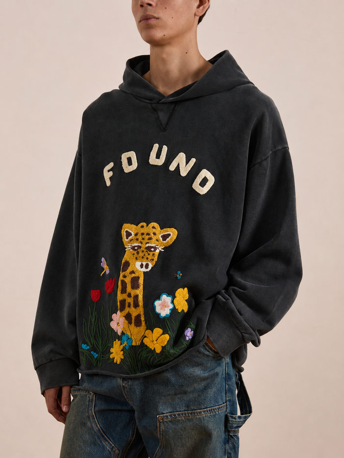 A person wearing the FOUND Embroidered Wildlife Hoodie—featuring “FOUND” text and an embroidered giraffe surrounded by flowers and butterflies on the front, crafted from vintage French-terry cotton—styled with blue jeans.