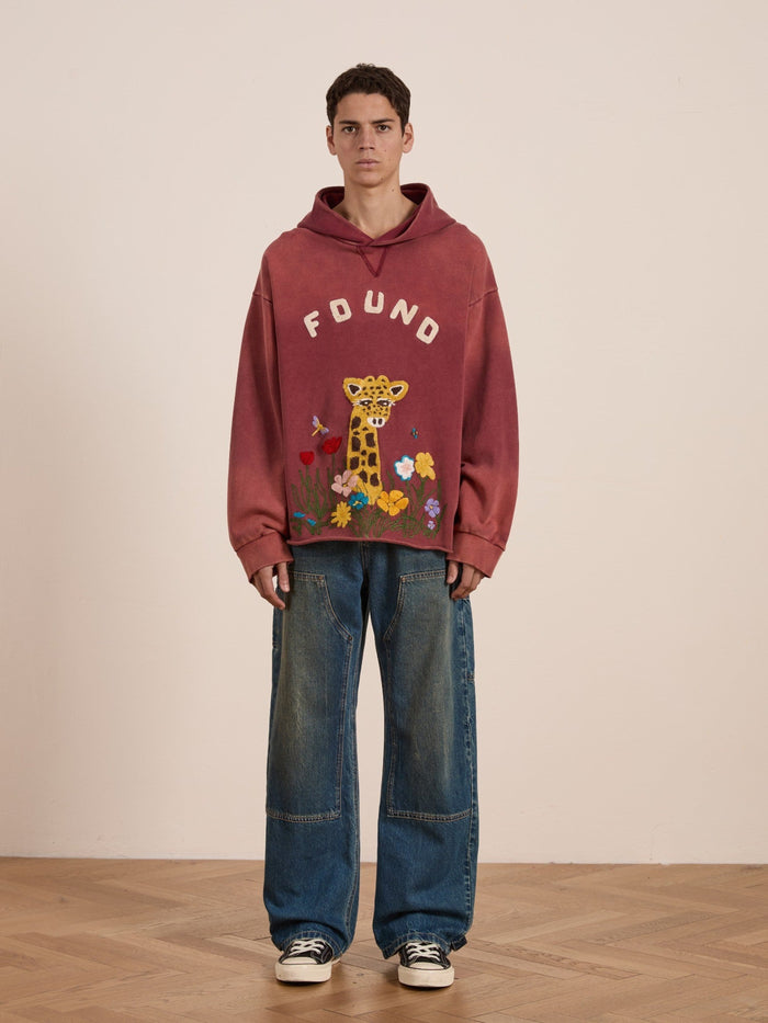 A young man stands indoors wearing the FOUND Embroidered Wildlife Hoodie with a giraffe design, styled with wide-leg jeans and black-and-white sneakers.