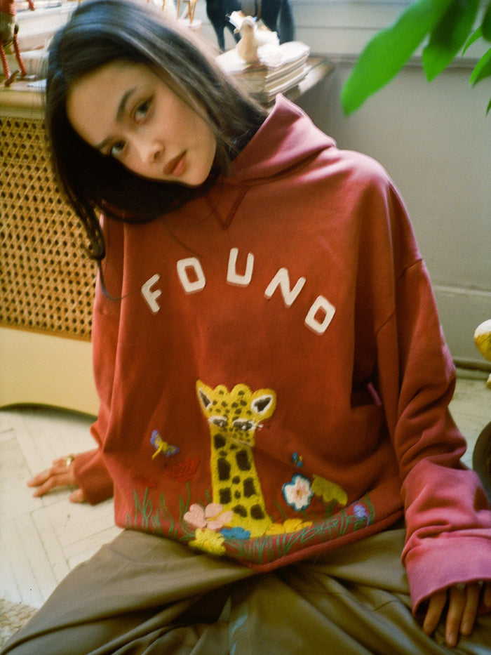 A woman sits indoors wearing the FOUND Embroidered Wildlife Hoodie, a red vintage French-terry cotton piece featuring "FOUND" and a floral giraffe embroidered on the front.