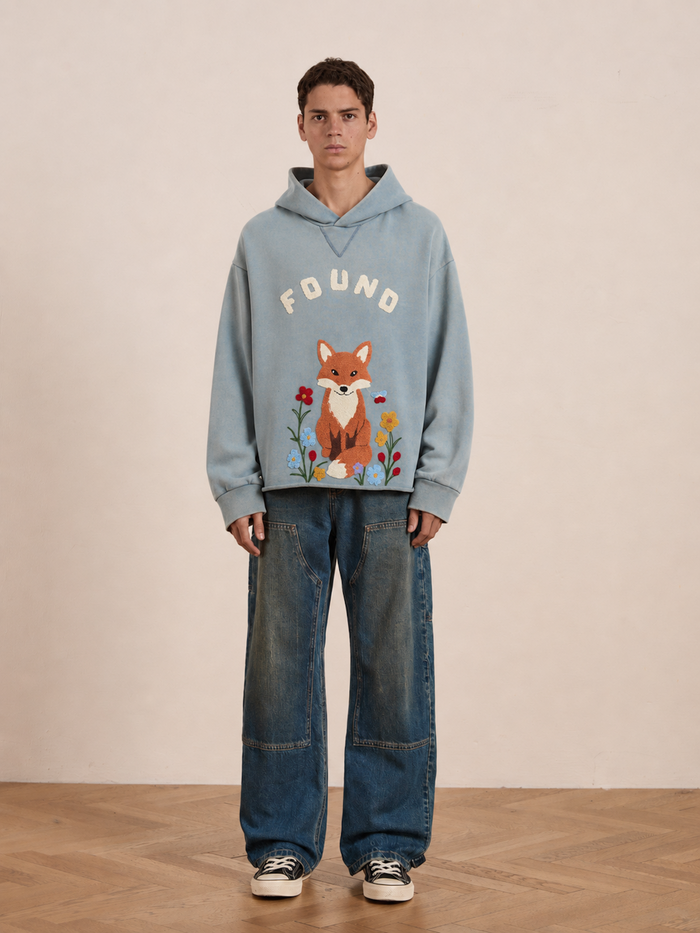 Indoors, a person stands against a plain background wearing the FOUND Embroidered Wildlife Hoodie—a blue, vintage French-terry hoodie featuring an embroidered fox and bold “FOUND” lettering—paired with baggy jeans and black-and-white sneakers.