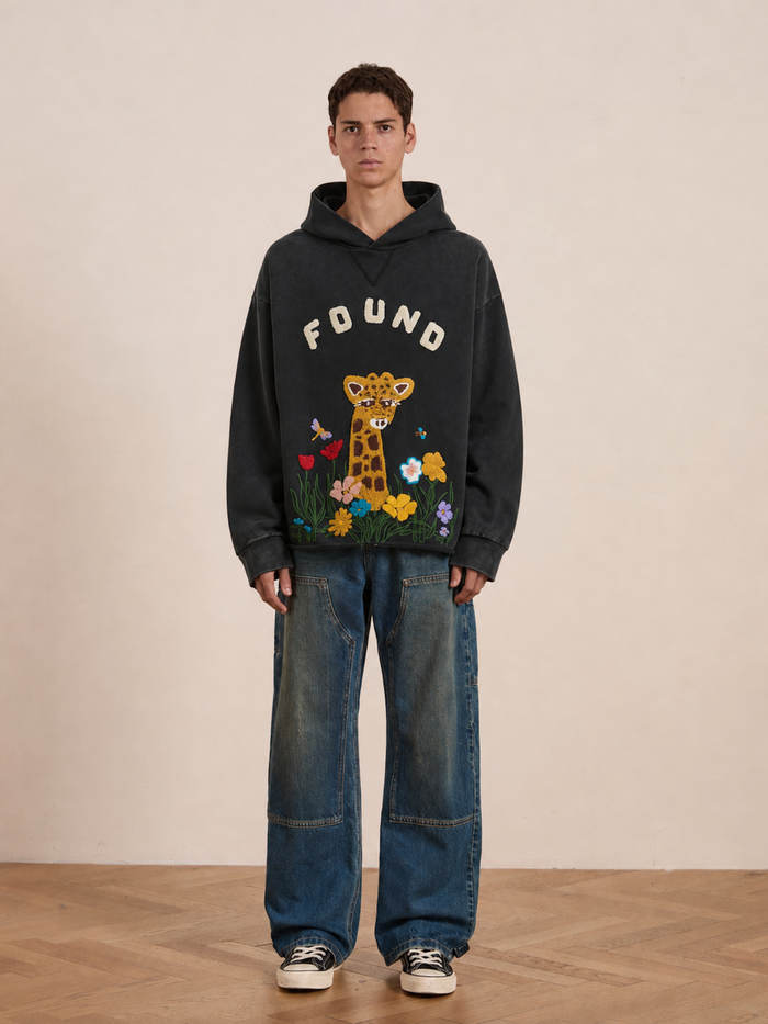 A person wears the FOUND Embroidered Wildlife Hoodie in black, featuring “FOUND” text and a cartoon giraffe amid flowers, crafted from vintage French-terry cotton. They pair it with baggy blue jeans and black sneakers, standing on a wooden floor against a plain wall.