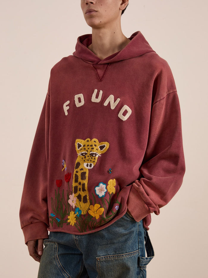 Someone wears a loose, faded red FOUND Embroidered Wildlife Hoodie in vintage French-terry cotton, featuring a giraffe with flowers and "FOUND" text on the front, paired with blue jeans.