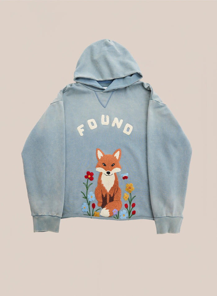 The Embroidered Wildlife Hoodie by FOUND is a light blue vintage French-terry hoodie featuring the brand’s “FOUND” text above a beautifully embroidered fox graphic sitting among flowers, shown on a plain background.