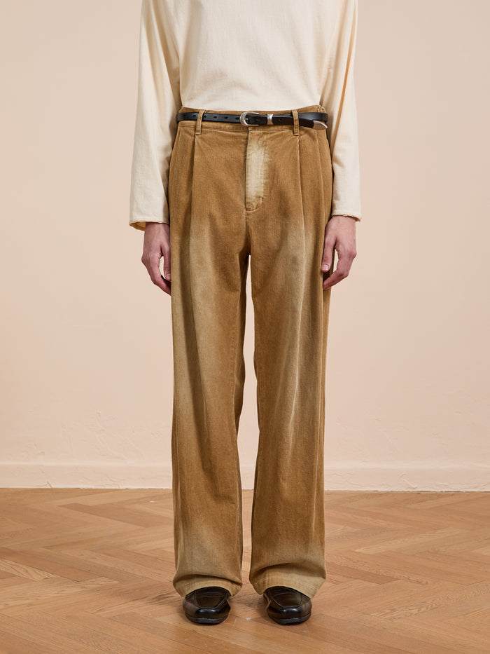 A person wears a cream long-sleeve shirt tucked into FOUND’s Faded Pleated Corduroy Pants with a black belt, standing on a wooden floor against a beige wall.