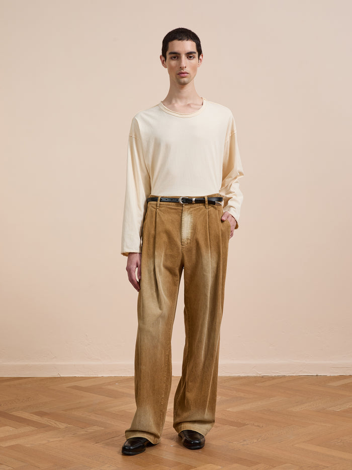 Indoors on a wooden floor, a person wears FOUND's Faded Pleated Corduroy Pants—high-waisted with double pleats and a relaxed fit—paired with a cream long-sleeve shirt, black belt, and black shoes.