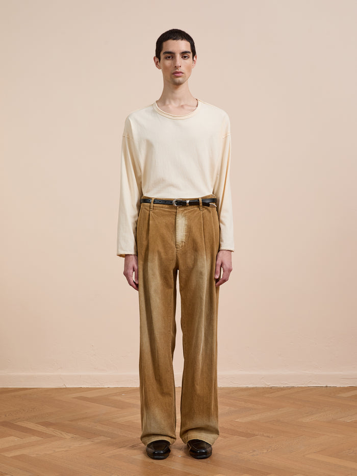 A person stands indoors against a beige wall, wearing a cream long-sleeve shirt, FOUND’s Faded Pleated Corduroy Pants, a black belt, and black shoes.