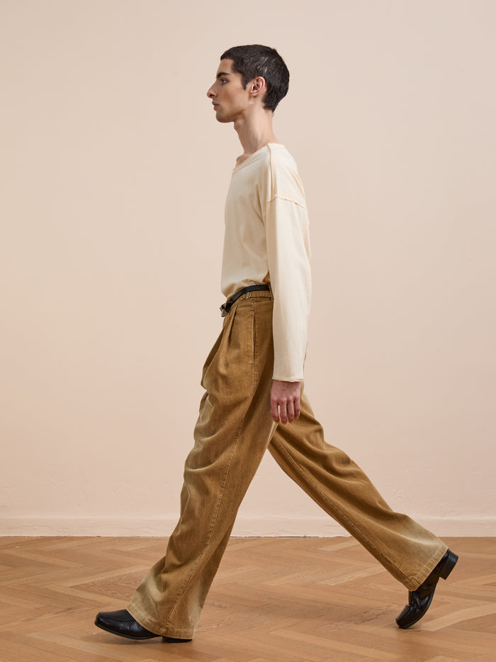 A person with short dark hair, in a cream long-sleeve shirt and FOUND's Faded Pleated Corduroy Pants, walks indoors on a wooden floor against a beige wall.