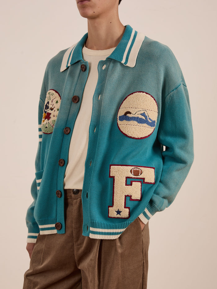 A person features the FOUND Fin Varsity Patch Collared Cardigan in teal, with prominent embroidered patches, over a crisp white shirt and classic brown pants, creating a collegiate style that radiates vintage charm.