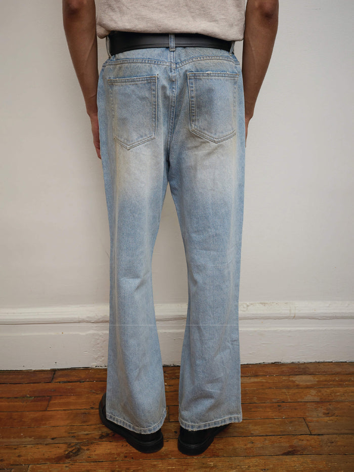 A person stands indoors on a wooden floor facing a wall, wearing Found's unisex light blue Flare Jeans and black shoes.
