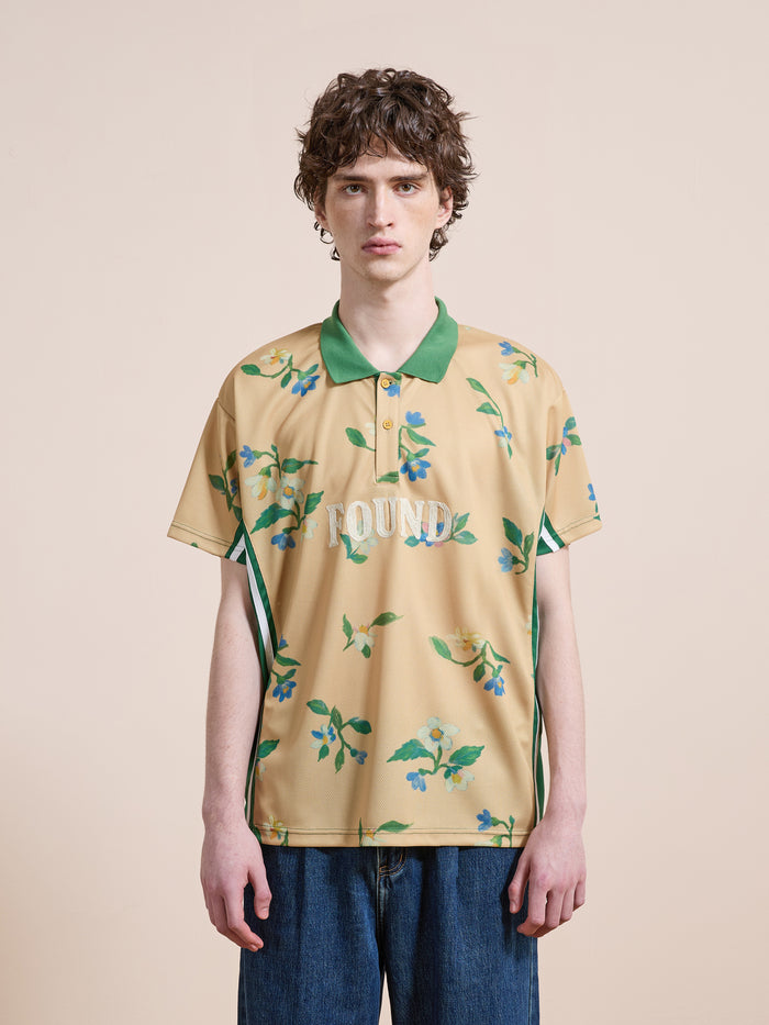 A person poses against a neutral backdrop in FOUND's Floral Polo Jersey—a tan floral polo with green collar and "FOUND" text—styled with blue jeans for a laid-back, vintage-inspired look.