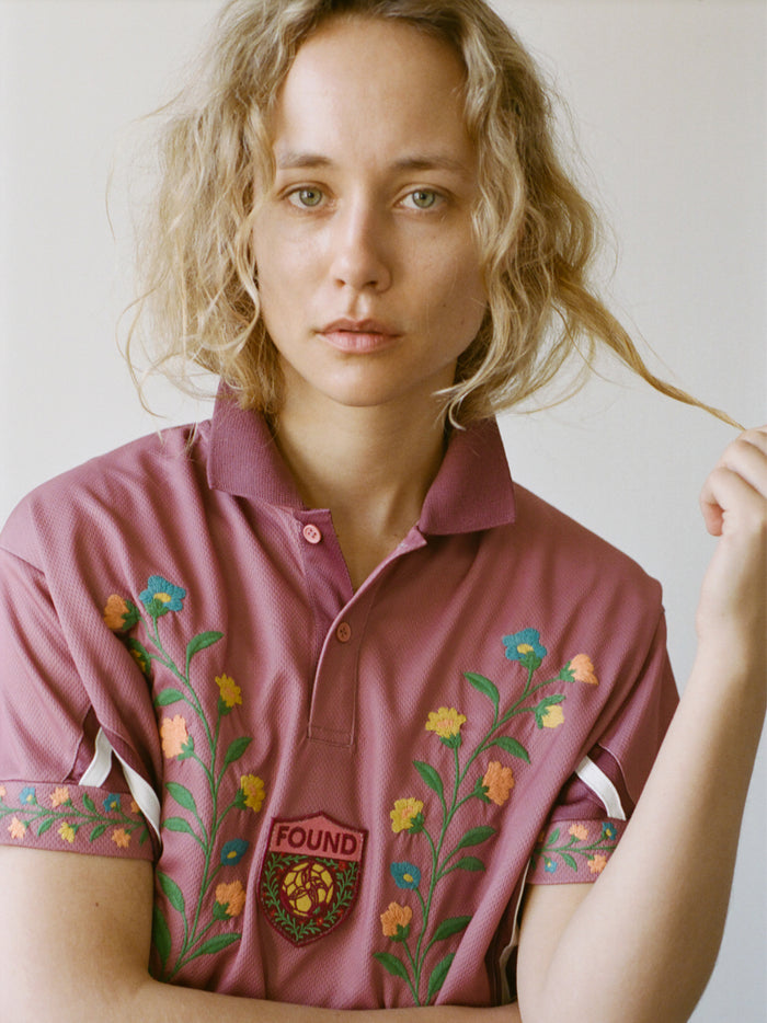 A person with wavy blonde hair wears the FOUND Floral Polo Jersey, a maroon cotton top featuring embroidered flowers and a "FOUND" patch, reminiscent of a vintage football shirt, and looks directly at the camera while holding a strand of hair.