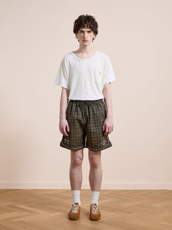 A young person stands indoors on a wooden floor, wearing Found's Forest Plaid Track Shorts with a white t-shirt, white socks, and brown shoes, facing the camera against a plain beige wall.