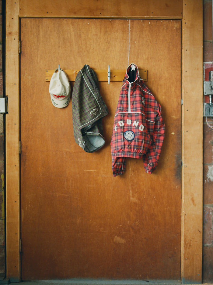 Beige cap, brown plaid shirt, and Found Forest Plaid Track Shorts with a unisex relaxed fit hang on hooks attached to a wooden door.