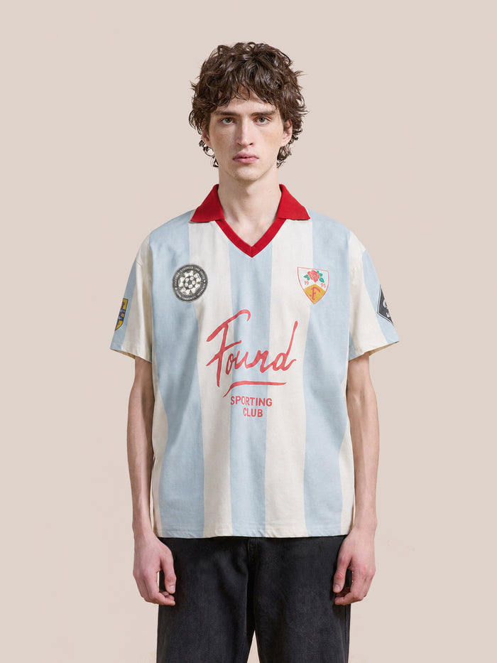 A person wearing the FOUND Found Club Short Sleeve Jersey—a unisex white and light blue striped top featuring a red collar, multiple patches, and “Found Sporting Club” text inspired by classic football shirts—stands against a plain background.