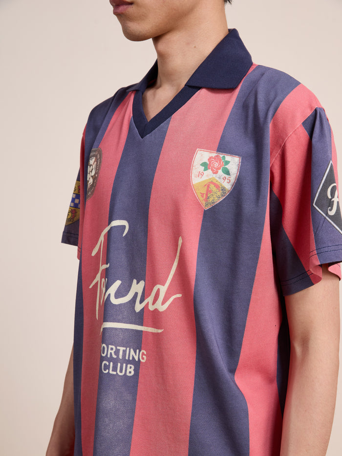 A person wearing the FOUND Found Club Short Sleeve Jersey—a unisex, relaxed fit vintage football shirt in blue and red stripes, featuring badges and “Ford Sporting Club” text on the front.