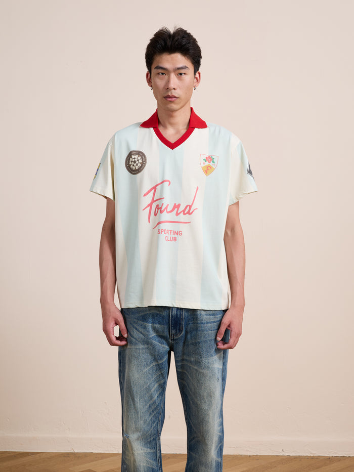 A person stands indoors against a plain wall, wearing the cream-colored FOUND Found Club Short Sleeve Jersey with jeans, facing the camera with a neutral expression. The unisex Found Club Short Sleeve Jersey by FOUND lends their outfit a classic sportswear vibe.
