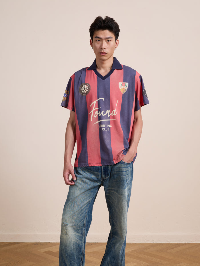 A person stands indoors against a plain background, wearing the FOUND Found Club Short Sleeve Jersey—a unisex vintage football shirt featuring pink and navy stripes—in a relaxed fit, paired with blue jeans and one hand in their pocket.