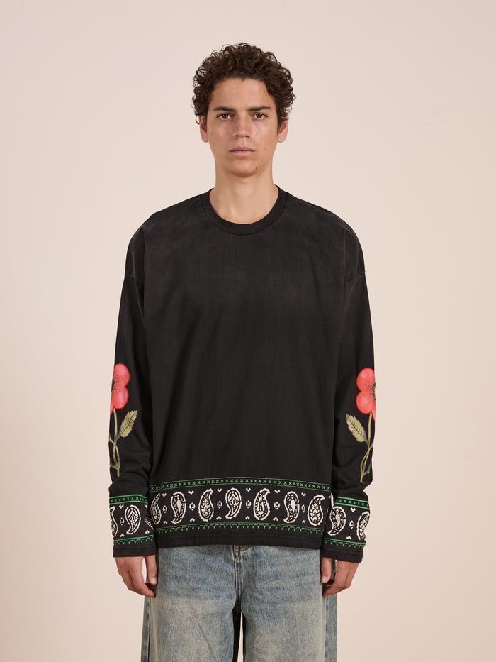 A person wears the FOUND Garden Bloom Embroidered LS Tee, a black long-sleeve with floral embroidery and paisley trim at the cuffs and hem, paired with light blue jeans, standing against a plain background in a relaxed fit.