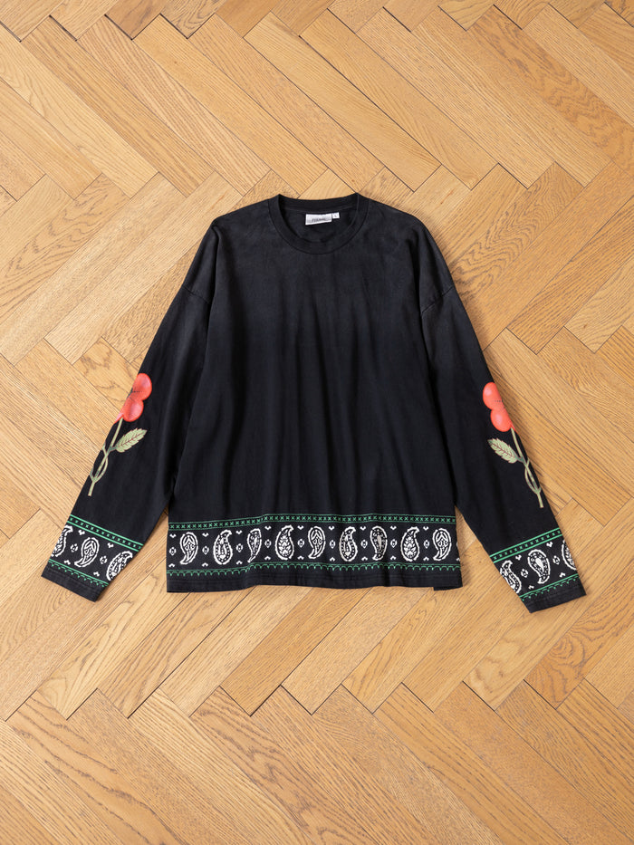 The FOUND Garden Bloom Embroidered LS Tee features floral embroidery on the sleeves, a white paisley pattern with green trim at the hem and cuffs, and a relaxed fit, shown laid flat on a wooden floor.