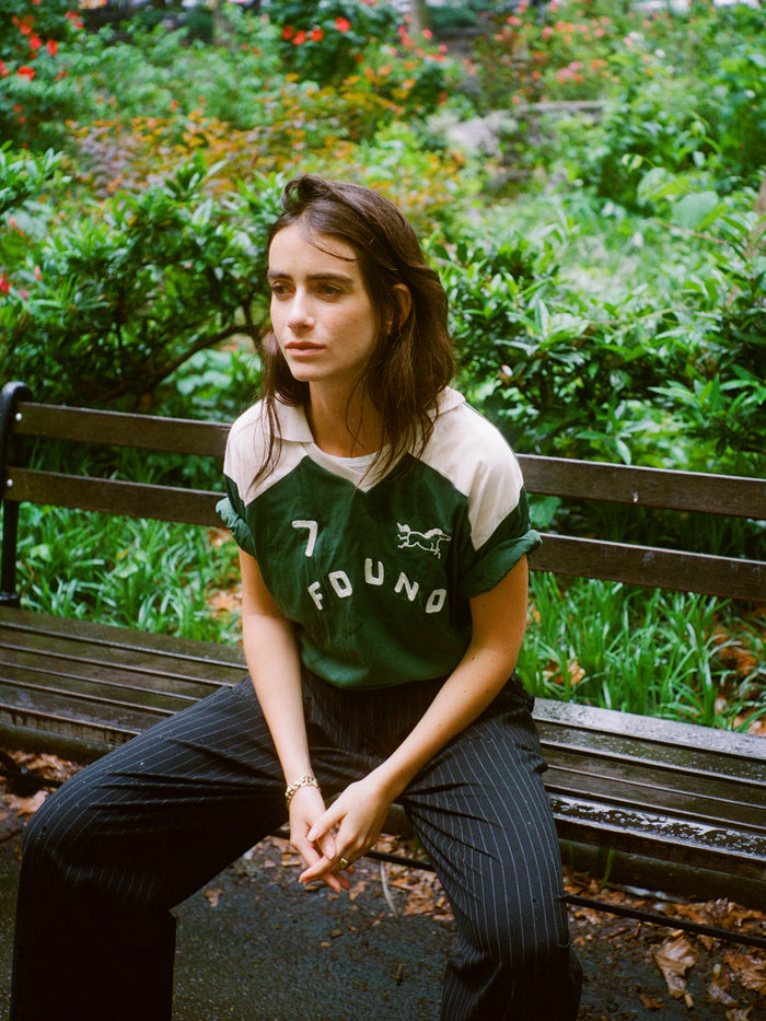 A woman with brown hair sits on a park bench, wearing the FOUND Goji Green Jersey and pinstripe pants. Green bushes and flowers fill the background.