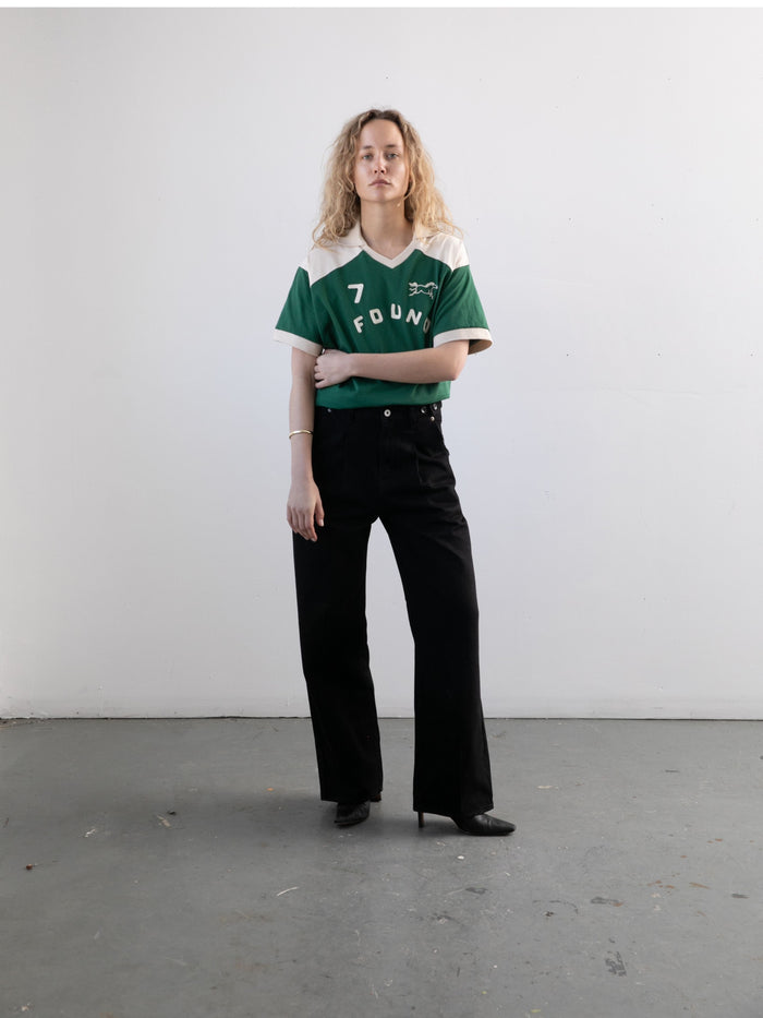 A person with curly hair stands indoors against a plain white wall, wearing the FOUND Goji Green Jersey, paired with black pants and black shoes.