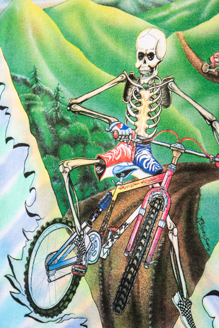 Skeleton riding a bicycle with colorful pants on a mountainous landscape