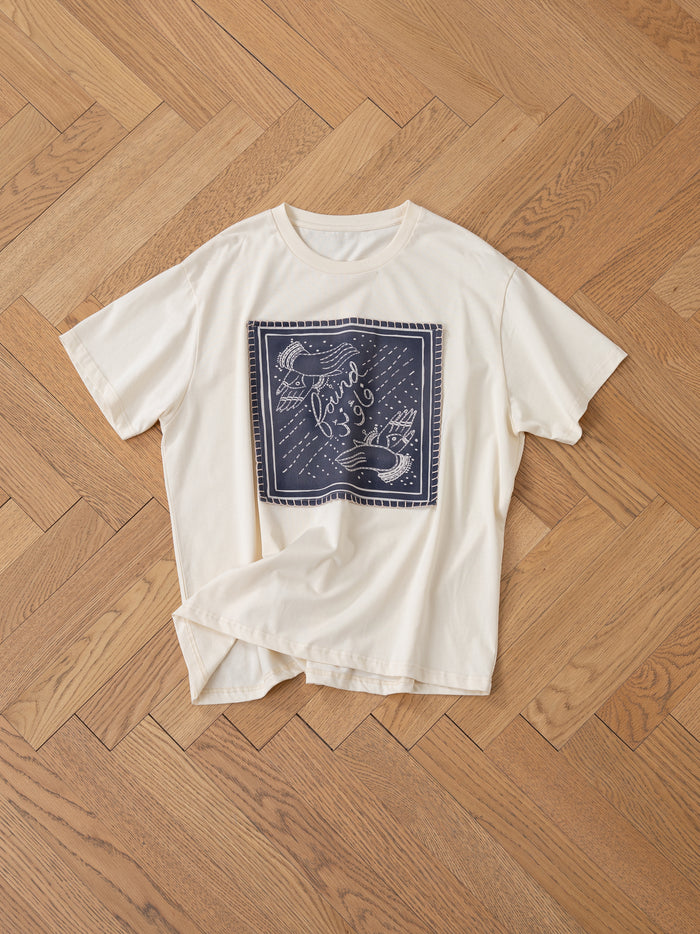 The Hand Art Graphic Tee by FOUND, a cotton cream t-shirt featuring a dark blue square graphic inspired by Mughal paintings with bird motifs and text, is elegantly laid on a wooden herringbone floor.