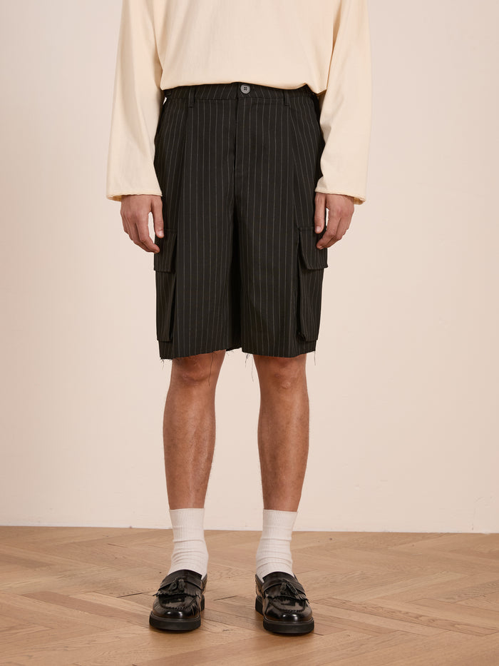 A person wears a cream long-sleeve shirt, FOUND Hava Pinstripe Pleated Cargo Trouser Shorts with an adjustable fit, white socks, and black loafers, standing on a wooden floor against a plain wall.