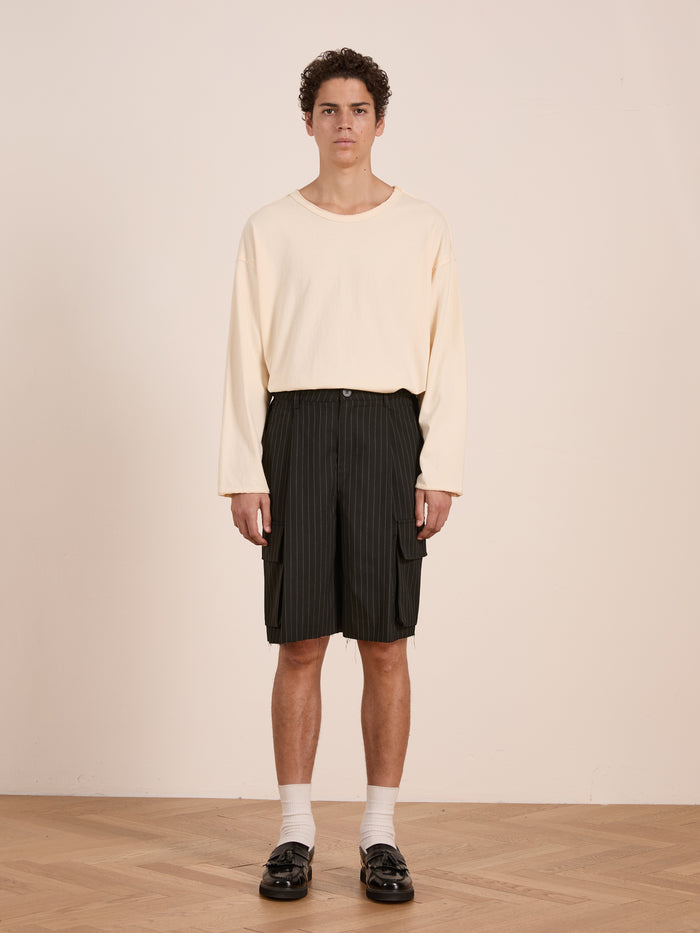 Indoors, a person wears a loose cream long-sleeve shirt, FOUND Hava Pinstripe Pleated Cargo Trouser Shorts, white socks, and black loafers on a wooden floor against a plain light-colored wall.