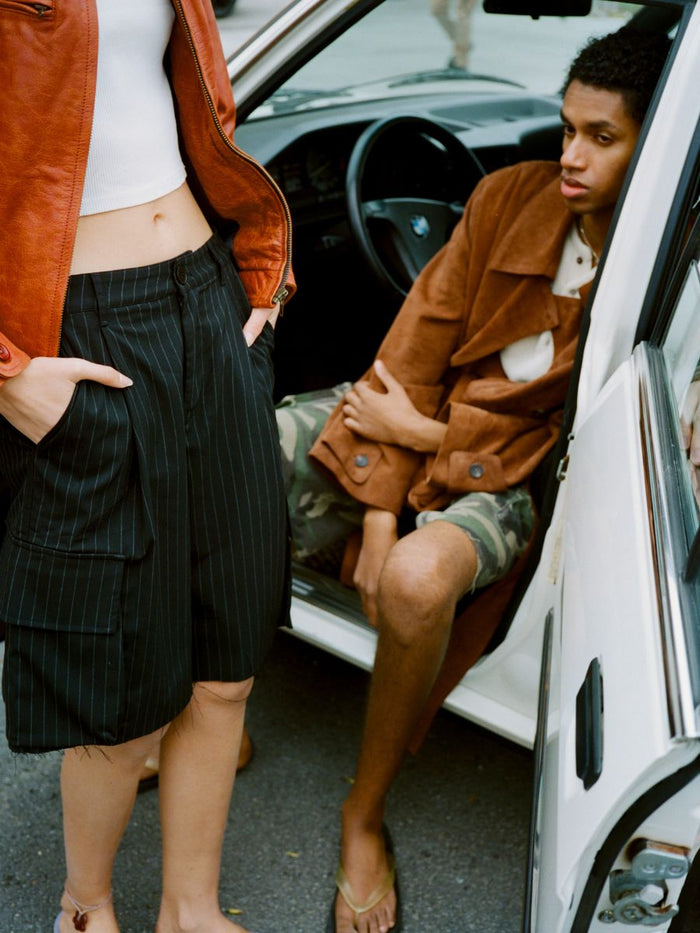 Two people stand by an open car door; one sits inside in a brown coat, while the other stands outside wearing a white top, brown jacket, and FOUND Hava Pinstripe Pleated Cargo Trouser Shorts.
