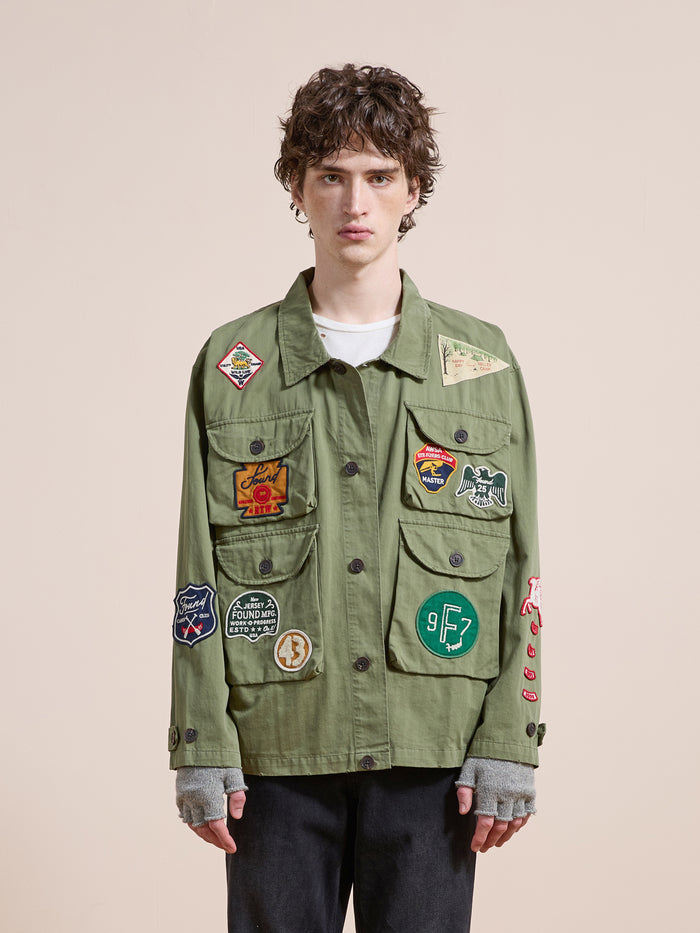 A young person with short, curly hair models the FOUND Herringbone Utility Patchwork Jacket—featuring colorful patches—with gray fingerless gloves and black pants, posed against a beige background.