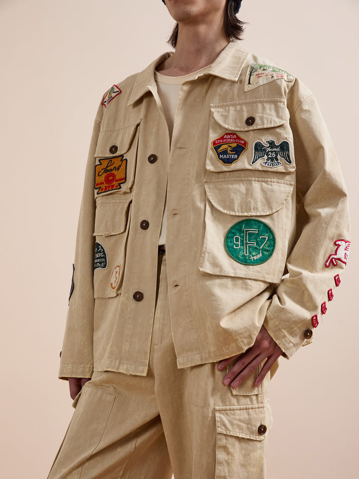 A person sports the FOUND Herringbone Utility Patchwork Jacket, featuring colorful embroidered patches on the chest, sleeves, and pockets, paired with matching pants against a plain background.
