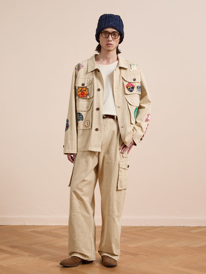 A person stands indoors on a wooden floor against a plain wall, wearing FOUND's Herringbone Utility Patchwork Jacket, matching cargo pants, white shirt, blue knit beanie, round glasses, and brown shoes.