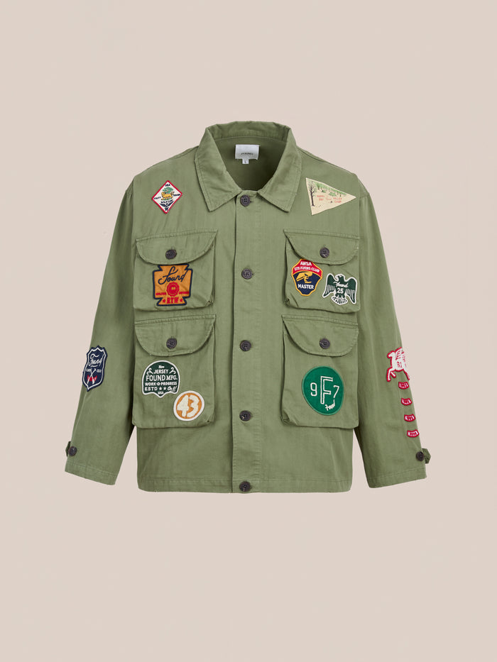 The FOUND Herringbone Utility Patchwork Jacket is a military-inspired olive green cotton jacket with multiple front pockets and colorful embroidered patches, shown against a plain beige background.