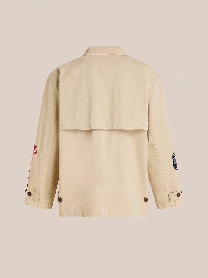 The FOUND Herringbone Utility Patchwork Jacket in beige is shown from the back, featuring long sleeves with buttoned cuffs and distinctive embroidered patches on each arm.