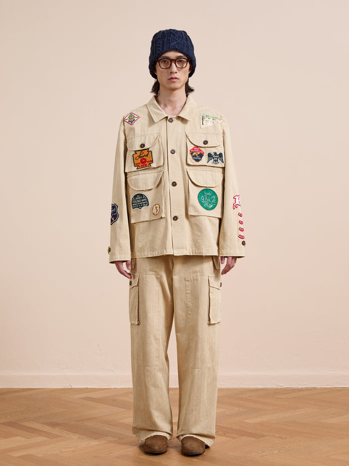 A person in glasses, a blue knit hat, and the FOUND Herringbone Utility Patchwork Jacket stands on a wooden floor against a plain, light-colored wall.