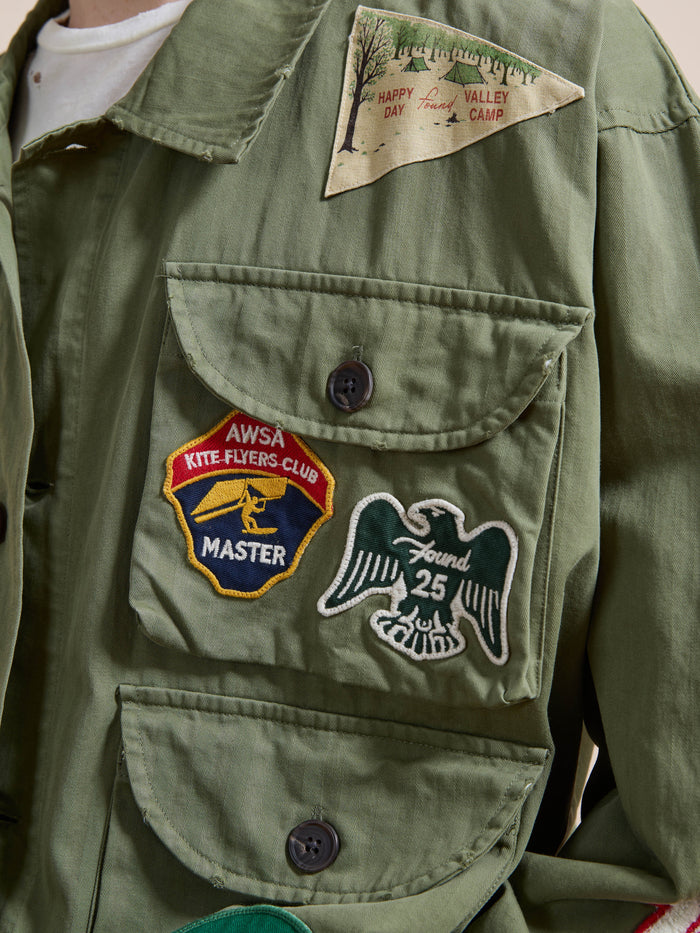 A person wears the FOUND Herringbone Utility Patchwork Jacket, featuring three embroidered patches: “AWSA Kite Flyers Club Master,” a bird with “Found 25,” and a triangle patch reading “Happy Valley Day Camp.”.