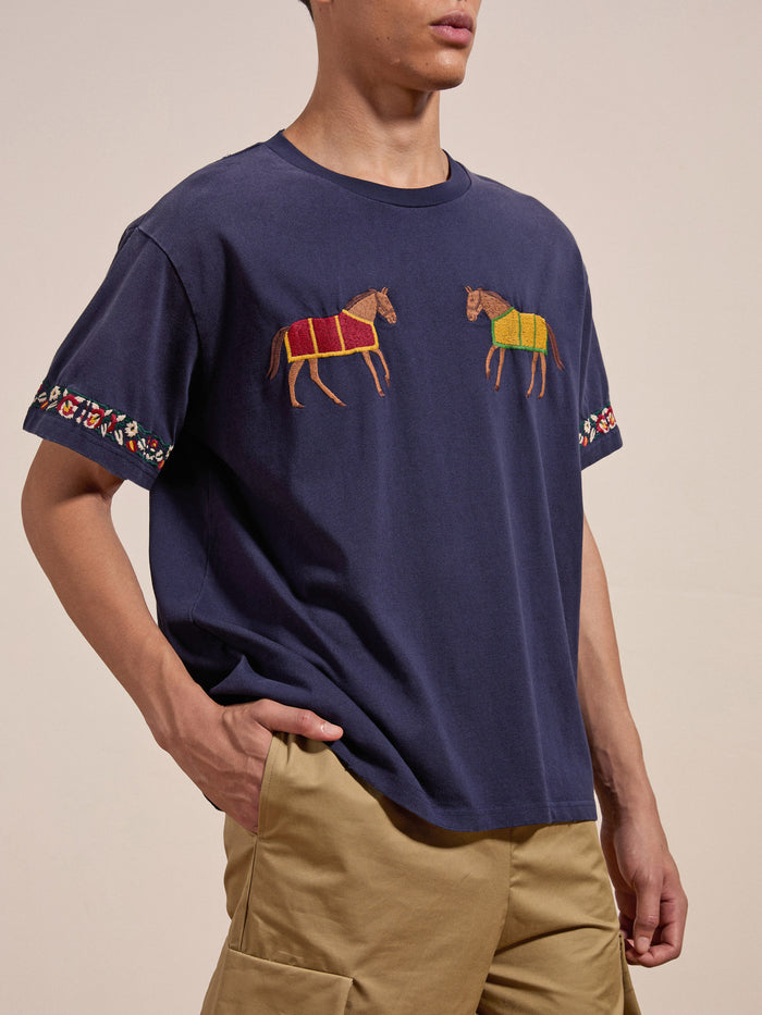 A person wearing the FOUND Horse Equine Tee, showcasing navy blue fabric with horse designs on the chest and vibrant embroidered patterns on the sleeves, paired with khaki pants.