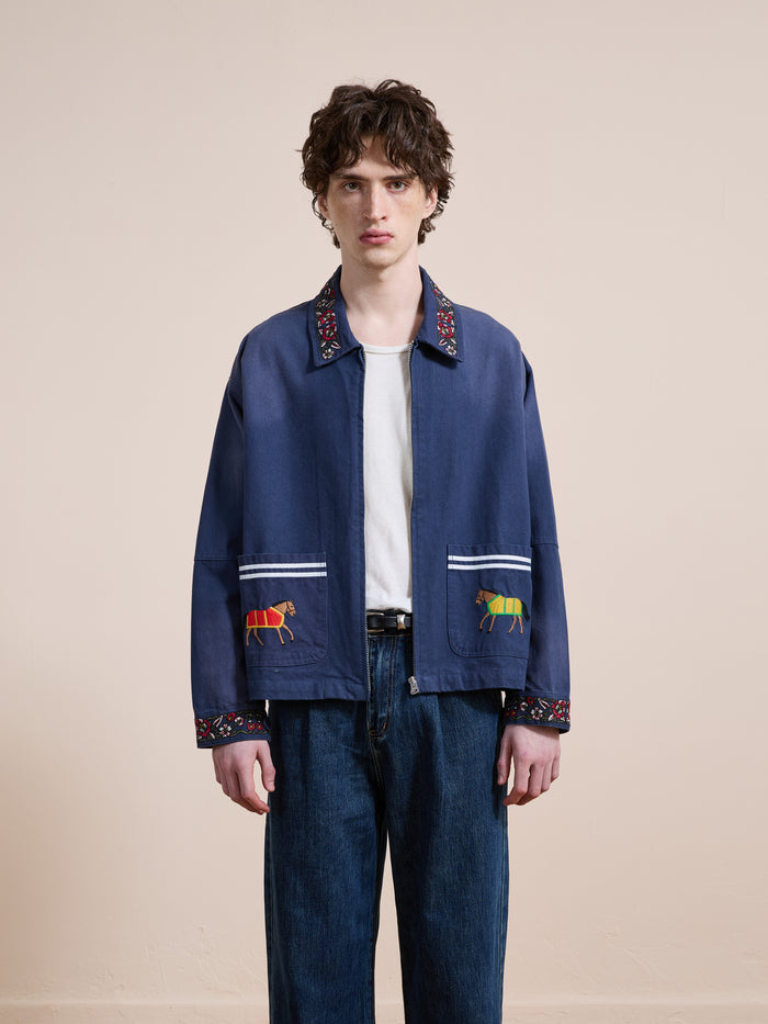 A young man faces forward, wearing the Found Horse Equine Work Jacket in cotton twill with animal-embroidered pockets, paired with a white t-shirt and blue jeans, set against a plain beige background.