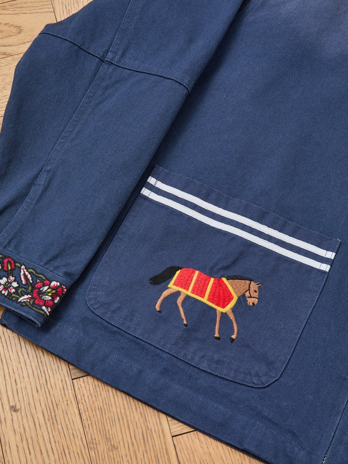 The Found Horse Equine Work Jacket in navy blue features a front pocket with an embroidered racing horse, white stripes above the pocket, and floral embroidery on the sleeve cuff.