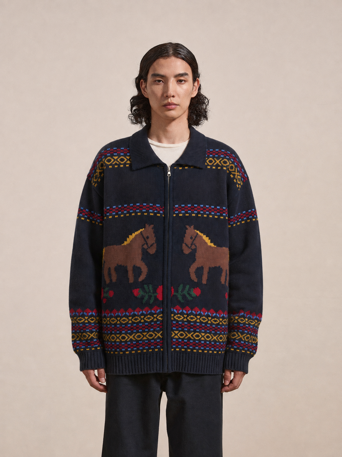 A person stands facing forward, wearing the Isles Sweater by Found—a navy knit zip cardigan detailed with jacquard horse motifs and floral patterns, accented by geometric designs in red, yellow, and green.