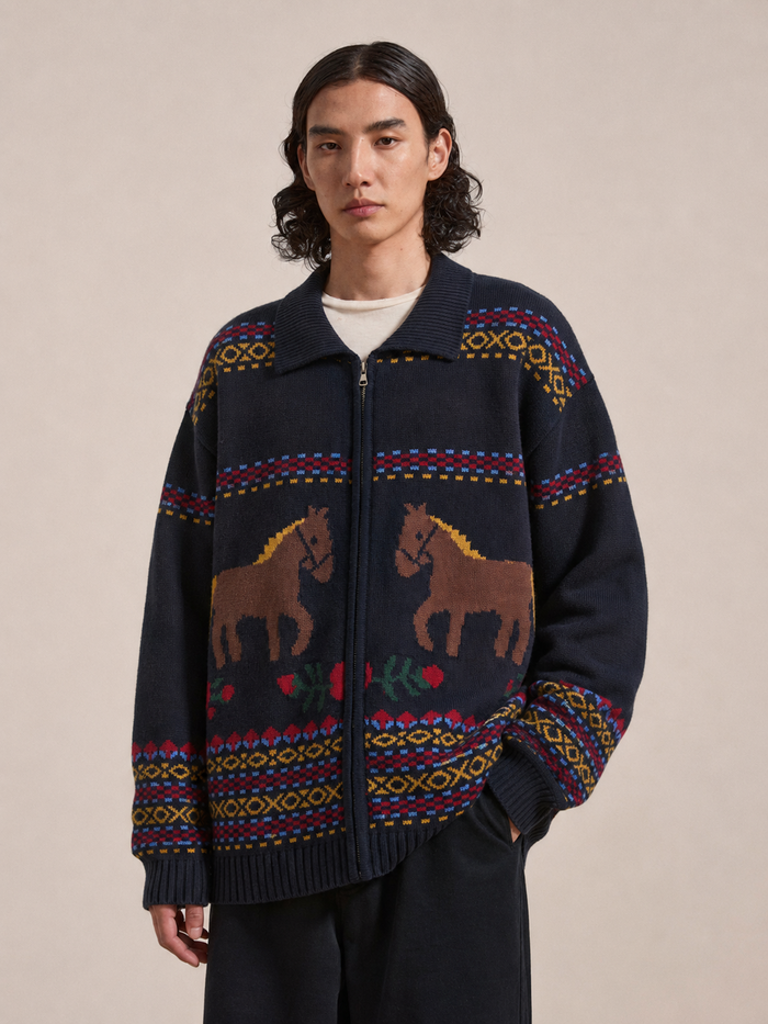 A person wearing the Found Isles Sweater—a navy knit zip cardigan adorned with colorful jacquard horse motifs—stands against a plain beige background.