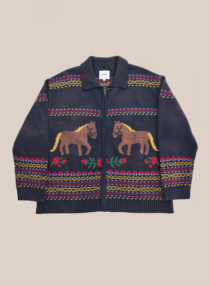 Introducing the Isles Sweater by Found: a navy blue knit zip cardigan featuring brown jacquard horse motifs, red floral patterns, and bold geometric designs on the sleeves and hem, finished with a ribbed collar and zip front.