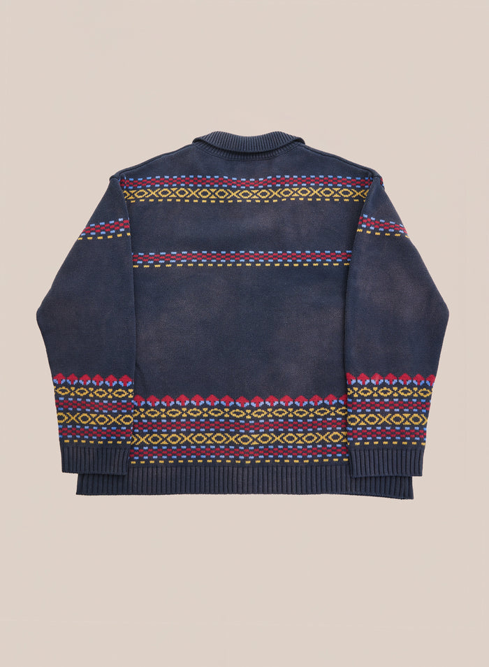 The Isles Sweater by Found, a navy knit featuring vibrant geometric patterns in yellow, pink, and green along the back, hem, and cuffs—plus understated jacquard horse motifs—is shown laid flat on a neutral backdrop.