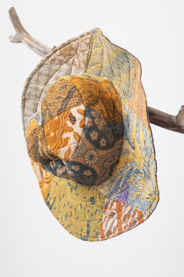 A reversible sun hat with a colorful geometric pattern on one side and a solid yellow color on the reverse, displayed on a piece of drift wood.