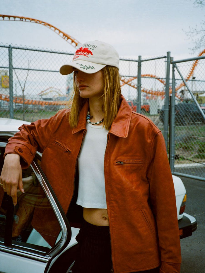 A person in a red leather jacket, white crop top, necklace, and the FOUND Jockey Club Found Cap stands by a white car; a rollercoaster and chain-link fence in the background add an equestrian touch.