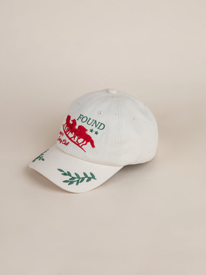 Introducing the Jockey Club Found Cap by FOUND: a stylish white cap adorned with intricate red embroidery of horses and the FOUND logo, complemented by green leaf designs on the brim.