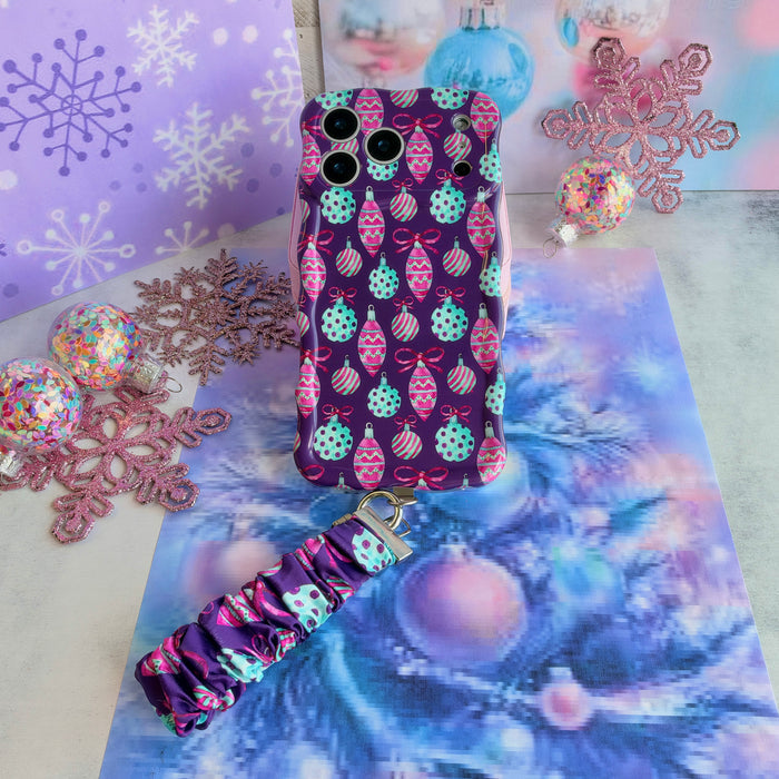 Just One More Ornament iPhone case with its ornament-print scrunchie wrist strap on lavender snowflake paper and aqua tinsel, beside a pink glitter snowflake