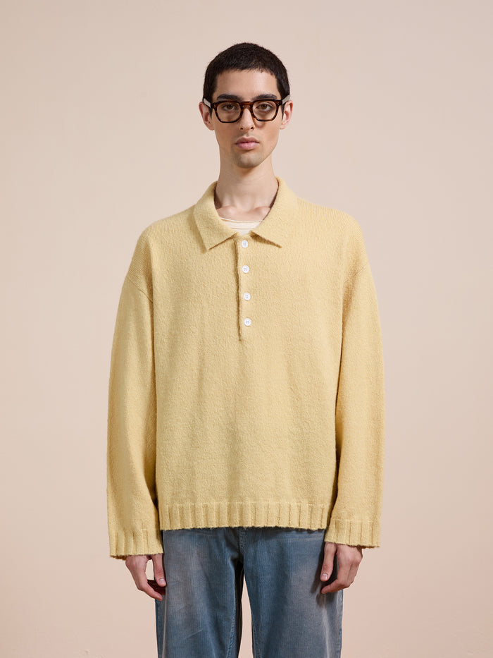 Someone wearing glasses, a pale yellow FOUND Knit LS Polo Sweater and loose blue jeans stands against a plain beige background.
